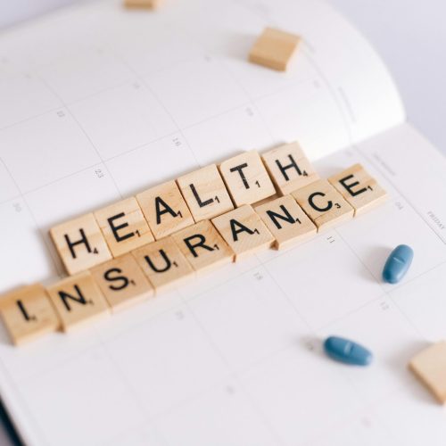 Close-up of health insurance spelled out with Scrabble tiles on a planner, alongside medication pills.